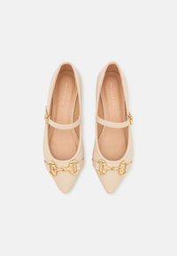 Ballerines Mary Jane beige à bout pointu avec détail mors doré et bride réglable, vues de dessus sur fond blanc.