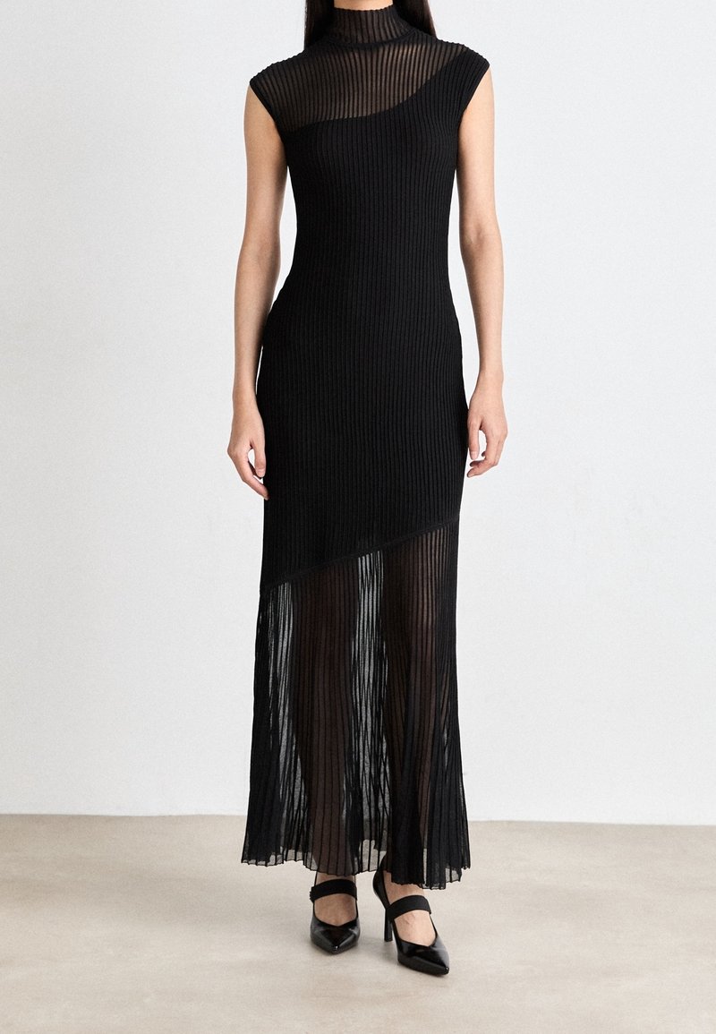 Femme portant une robe noire sans manches côtelée avec un tissu transparent sur la partie inférieure et la poitrine, associée à des talons noirs à bout pointu.