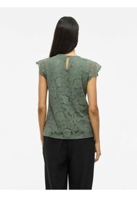 Kanten blouse in gedempt groen met een bloemenpatroon, korte franje mouwen en een keyhole detail op de rug. G gedragen met zwarte broeken.