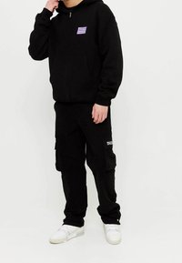 Schwarze Zip-up-Hoodie mit Fronttaschen und einem kleinen lila-weißen Grafik, kombiniert mit schwarzen Cargo-Hosen mit Seitentaschen und Text.