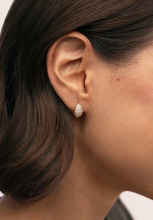 Pendiente de lágrima de oro con superficie texturizada y strass transparentes, mostrado en un primer plano junto a una oreja y un hombro.