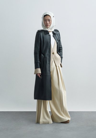 Μαύρο δερμάτινο παλτό, μπεζ oversized παντελόνι, και κρεμ πλεκτή κουκούλα. Υφασμάτινη υφή, δομημένη σιλουέτα και λεπτομέρεια κουμπιού ορατή.