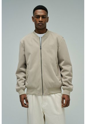 Homme portant une veste bomber beige à fermeture éclair et un pantalon blanc, debout devant un fond gris uni.