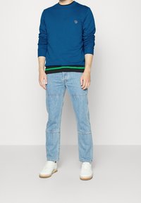 Blauwe sweatshirt met een ronde hals en lange mouwen, voorzien van een klein logo. Gecombineerd met lichtblauwe spijkerbroek en witte sneakers.