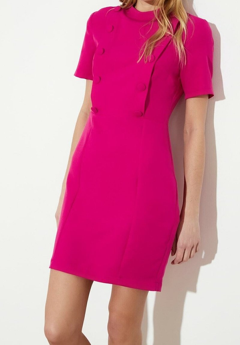 Femme portant une robe rose vif à manches courtes, longueur genou, avec des boutons recouverts de tissu sur le devant, sur fond blanc.