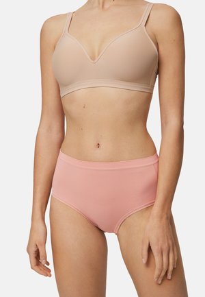 Mujer con un sostén beige sin costuras y ropa interior rosa de talle alto, de pie contra un fondo blanco liso.