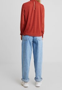 Rote gemusterte Bluse mit hohem Kragen und gerafftem Rücken, kombiniert mit lockeren blauen Jeans, die an den Säumen umgeschlagen sind, getragen mit weißen Sneakers.