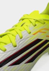 Λευκό και φωτεινό κίτρινο ποδοσφαιρικό παπούτσι με μαύρες ρίγες, υφή στην επιφάνεια και ζωντανά κορδόνια. Διαθέτει αεροδυναμικό σχεδιασμό και αντιθετικές λεπτομέρειες.