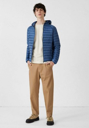 Giovane uomo in piedi con le mani nelle tasche, indossa una giacca a cappuccio imbottita blu, una maglietta crema, pantaloni color sabbia e sneakers color sabbia con suole nere.