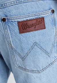 Jeans de mezclilla azul claro con una etiqueta de cuero marrón "Wrangler" en el bolsillo trasero, con costuras en contraste y un patrón de costura distintivo.
