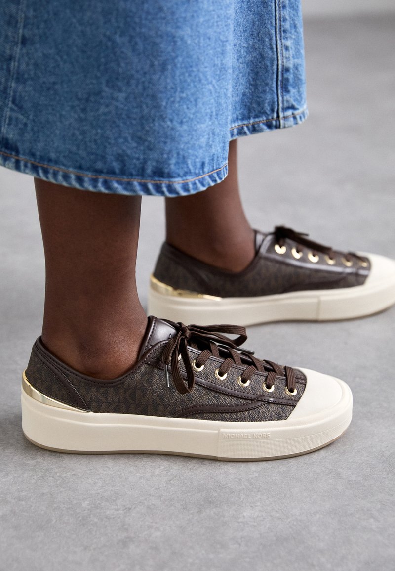 Brown canvas sneakers met een monogram patroon, voorzien van gouden hardware, beige zolen en bruine veters. G gedragen met een spijkerrok.