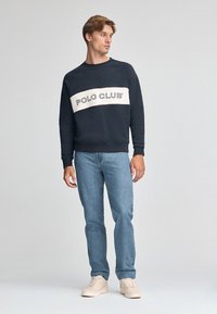 Felpa blu navy con logo a strisce bianche "POLO CLUB" sul petto, abbinata a jeans azzurri chiari e sneakers beige. Materiale misto cotone.