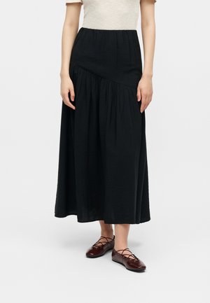 Kvinde iført en sort asymmetrisk midinederdel med rynkede detaljer, beige top og mørkerøde snørelace flats på hvid baggrund.