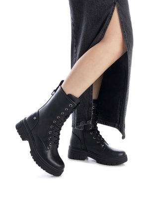 Stivali alla caviglia in pelle nera con design allacciato, punta rotonda, suola spessa e occhielli metallici. Indossati con una gonna di jeans nera con spacco alto.