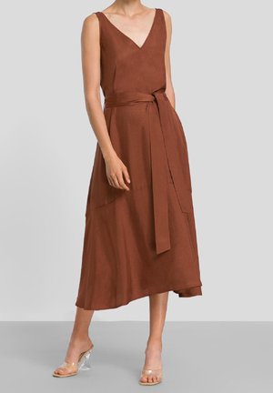 Ærmeløs brun midi-kjole med V-udskæring og taljesnor, båret med transparente hælede sandaler på en ensartet grå baggrund.