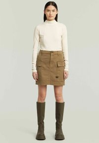 Maglietta beige a maniche lunghe con collo alto, abbinata a una gonna cargo khaki con tasche anteriori e patch del brand. Stivali alti fino al ginocchio verde oliva.