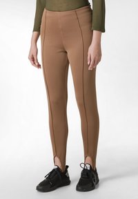 Hnědé legíny s hladkou, elastickou texturou, obsahující postranní švy a rozparky na kotnících, spárované s černými sportovními botami.