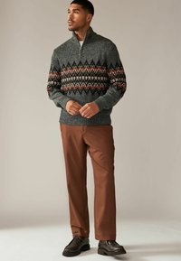 Pull en maille gris foncé avec un col zippé, arborant un motif géométrique coloré. Un pantalon marron et des chaussures foncées complètent la tenue.