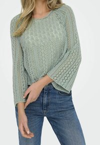 Pull vert clair en maille avec un motif texturé, manches larges et encolure ronde, associé à un jean bleu.