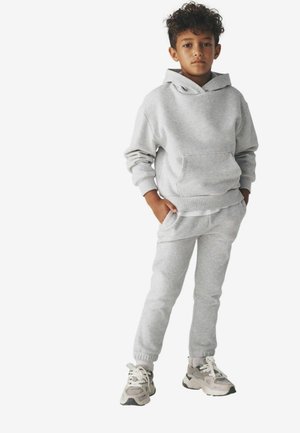 Chłopak z kręconymi włosami stoi z rękami w kieszeniach, ubrany w jasnoszary hoodie, pasujące dresy i białe trampki na białym tle.