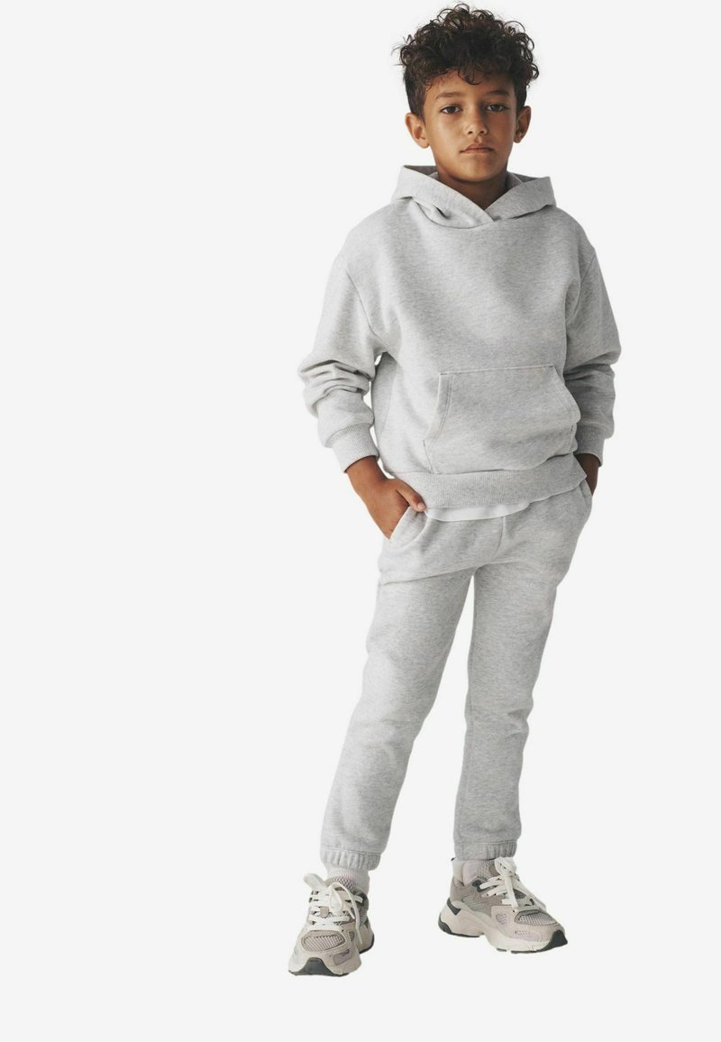 Chłopak z kręconymi włosami stoi z rękami w kieszeniach, ubrany w jasnoszary hoodie, pasujące dresy i białe trampki na białym tle.
