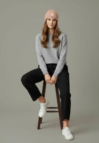Gray sweater, black pants, pink ribbed beanie with logo, and white sneakers. The model sits on a stool against a neutral background.