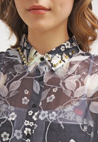 Transparente Bluse mit floralem Muster auf dunklem Hintergrund, mit goldenen und silbernen Nieten am Kragen, schwarzen Knöpfen und detaillierten botanischen Motiven.