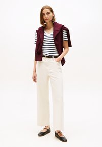 Jauna sieviete valkā baltas platas džinsu bikses, melnas kurpes ar zemu papēdi, svītrainu V veida kakla kreklu un tumši sarkanu džemperi, kas pārmests pār pleciem, stāv ar roku kabatā.