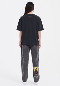 Př oversized černém bavlněném tričku s červeným logem na zádech, které je spojeno s vybledlými šedými džíny s barevným vlčím grafickým motivem na nohavici.