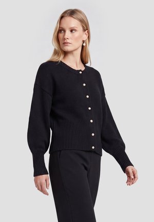 Lyshåret kvinde iført sort strikket cardigan med perleknapper og matchende sorte bukser, som kigger til venstre mod en ensfarvet baggrund.