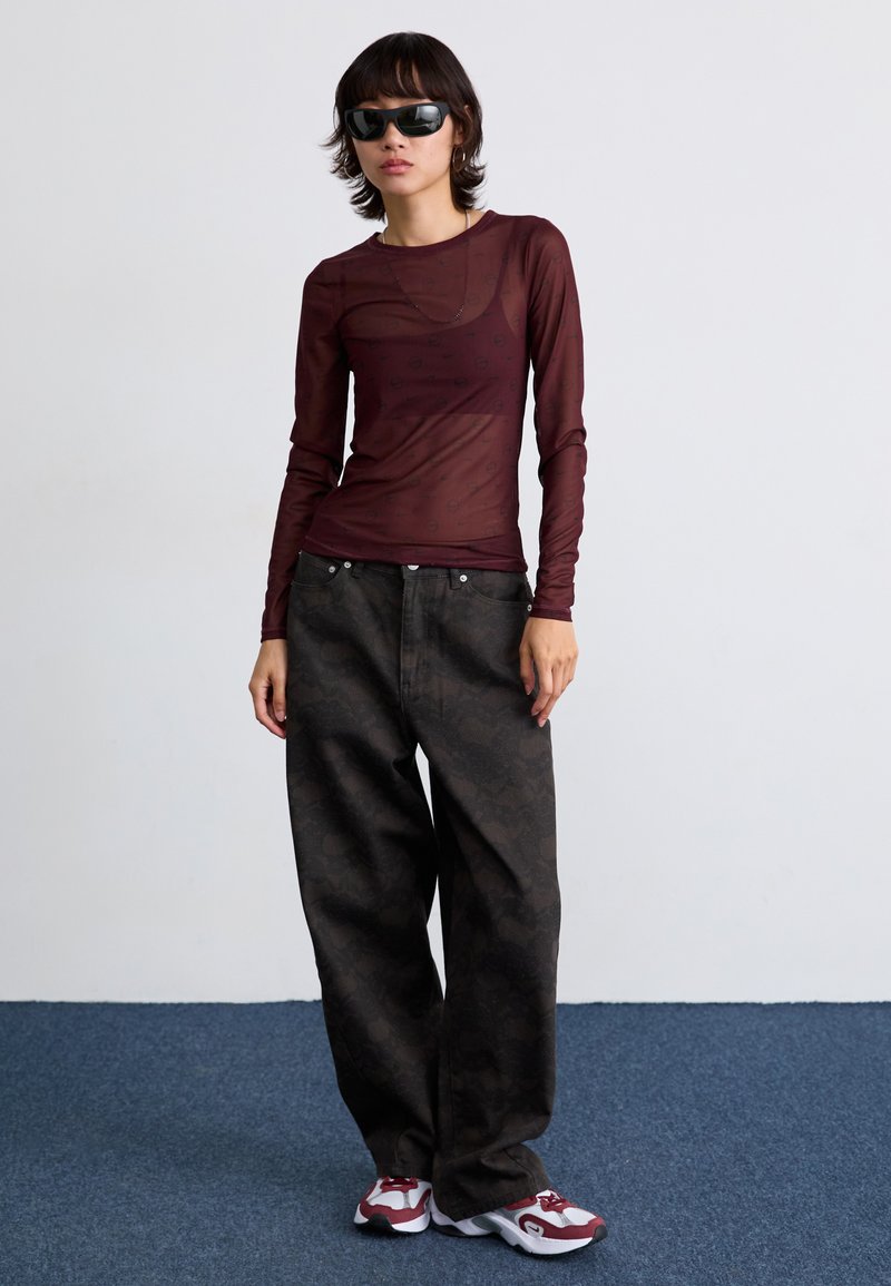 Top transparente de manga larga en color burdeos con patrón, combinado con pantalones de pierna ancha de patrón oscuro. Unas gafas de sol negras y zapatillas estilizadas completan el look.