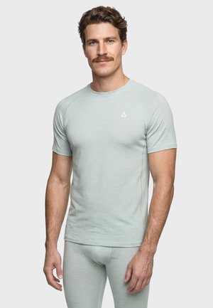 Man with brown hair and mustache wearing light gray fitted short-sleeve shirt and matching pants, standing against plain light background.