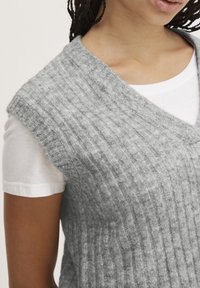 Grau, gerippter Strickweste mit V-Ausschnitt und ärmellosem Design, kombiniert mit einem weißen T-Shirt, das eine strukturierte Oberfläche aufweist.