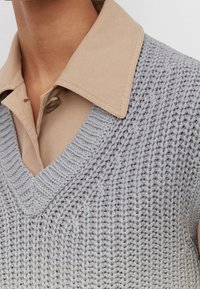 Gilet tricoté gris avec col en V, présentant un motif tissé texturé, superposé à une chemise col beige clair. Boutons visibles sur la chemise.