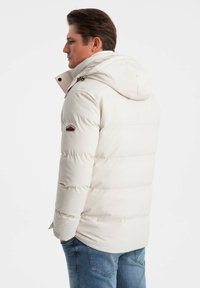 Giacca puffy bianca con cappuccio, caratterizzata da cuciture orizzontali e una patch con logo sulla manica sinistra. Indossata con jeans blu.