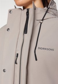 Veste beige clair avec un col montant, une fermeture éclair et une capuche à cordon de serrage. Caractérisée par des éléments noirs et le nom de la marque "DIDRIKSONS" imprimé.