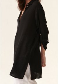 Camicia nera con colletto, realizzata in tessuto testurizzato, dotata di maniche lunghe, scollatura a V profonda e spacchi laterali per una vestibilità rilassata. Bottoni dorati accentuano il colletto.