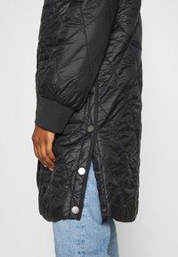 Giacca trapuntata nera con chiusura a zip, bottoni a pressione, polsini elastici e design fino al ginocchio realizzata in materiale sintetico lucido.