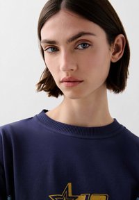 Sudadera navy con cuello de canalé, que presenta un diseño de estrella dorada brillante. Textura de tela suave, usada por una persona con el cabello corto.