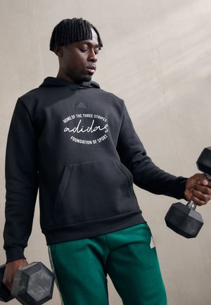 Sudadera negra con capucha y texto blanco. El modelo sostiene mancuernas, combinada con pantalones deportivos verdes con rayas blancas. Tela suave, corte relajado.