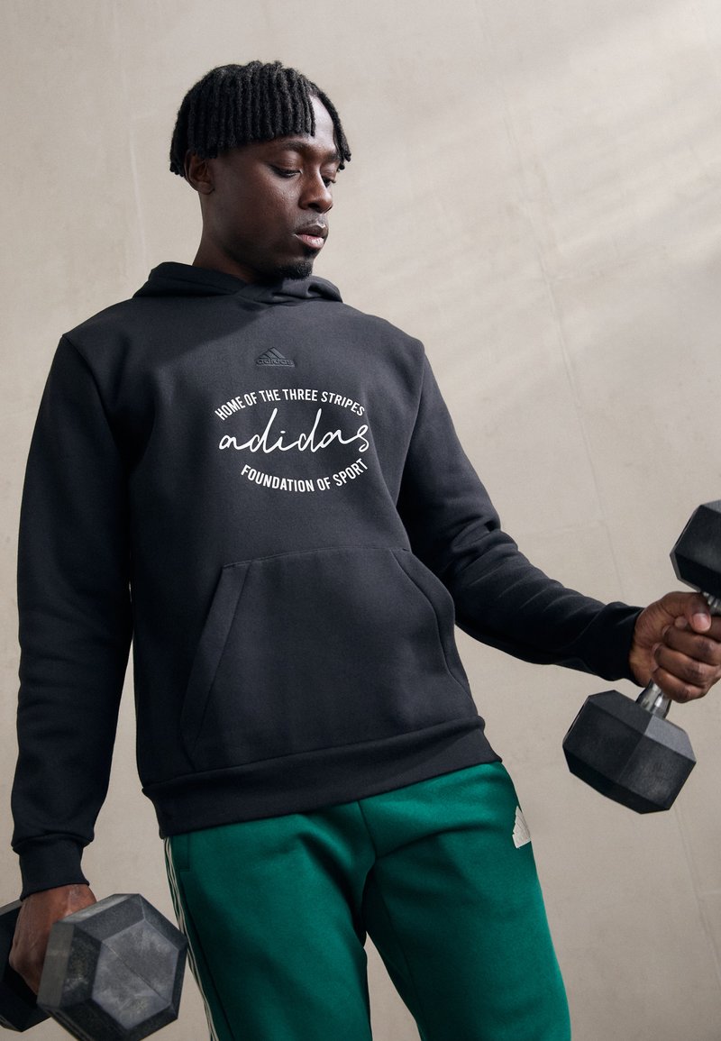 Felpa con cappuccio nera con testo bianco. Il modello tiene dei manubri, abbinata a pantaloni sportivi verdi con strisce bianche. Tessuto morbido, vestibilità rilassata.