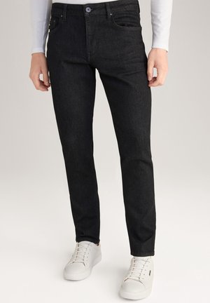 Person trägt schwarze Slim-Fit-Jeans mit Taschen und weiße Schnürer-Sneaker und steht vor hellem Hintergrund.