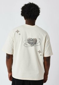 Camiseta de manga corta color crema con un gráfico de corazón y texto en espiral "PROYECTO PARÍS", acentuado por formas de estrellas en tonos apagados.