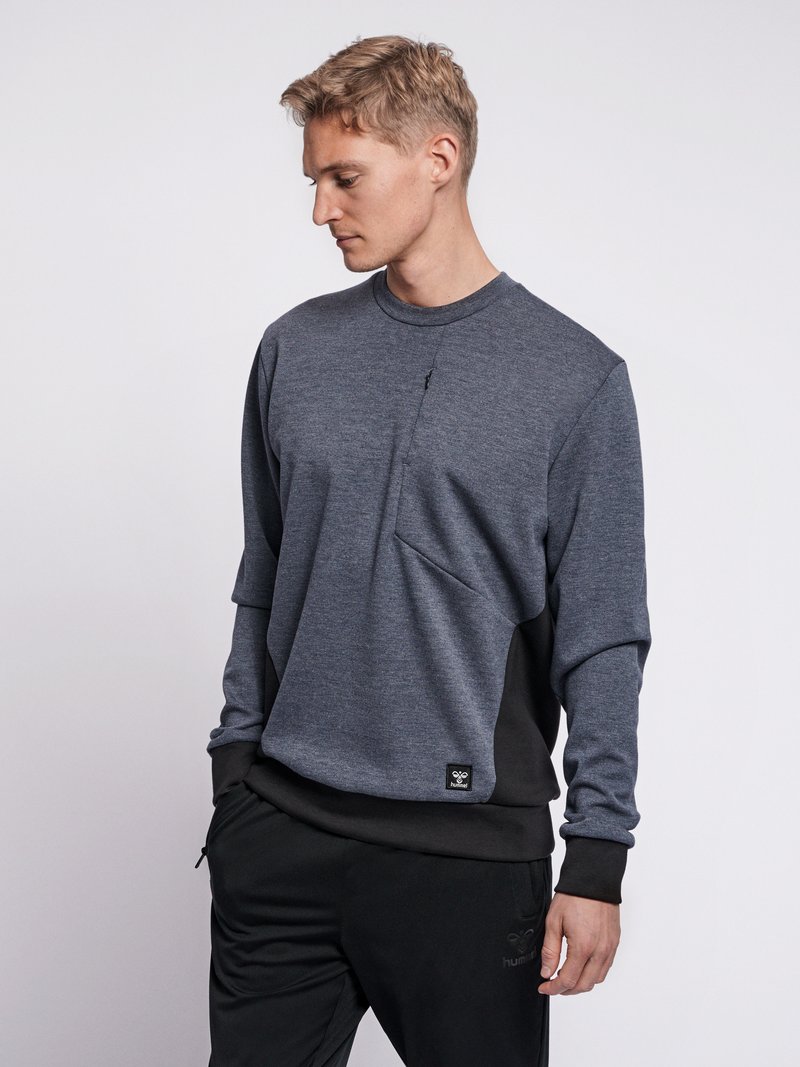 Ung mand iført en tofarvet grå og sort sweatshirt og sorte bukser, kigger ned og til venstre med hænderne i lommerne.