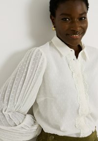 Camisa blanca de manga larga con tejido texturizado, mangas abullonadas con pliegues, borde de encaje a lo largo de la tapeta de botones y botones redondos.