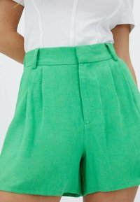 Pantaloni corti verdi a vita alta realizzati in un tessuto texturizzato, caratterizzati da pieghe frontali e tasche laterali per una maggiore funzionalità.