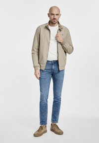 Kurtka bomber w kolorze tan, lekka, z ściągaczami, pod nią biała koszulka, niebieskie dżinsy i brązowe sneakersy. Prosty wzór, casualowy styl.