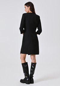 Frau in einem schwarzen knielangen Kleid mit Dreiviertelärmeln und schwarzen kniehohen Lederstiefeln, die vor einem einfarbigen Hintergrund steht.