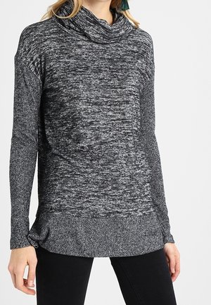 Femme portant un pull en maille gris chiné à manches longues avec un col bénitier ample et un pantalon noir.