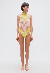 Γυναίκα που στέκεται ξυπόλητη φορώντας μια αμάνικη κορμά με υψηλό λαιμό σε κίτρινο και ροζ διακοσμητικό σχέδιο, κοιτάζοντας πλάγια σε απλό φόντο.